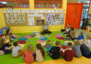 Grupa dzieci siedzi w półkolu wokół pani Magdaleny Siupy dyrektor Miejsko- Gminnej Biblioteki w Kleczewie, która prezentuje książki o tematyce świątecznej.